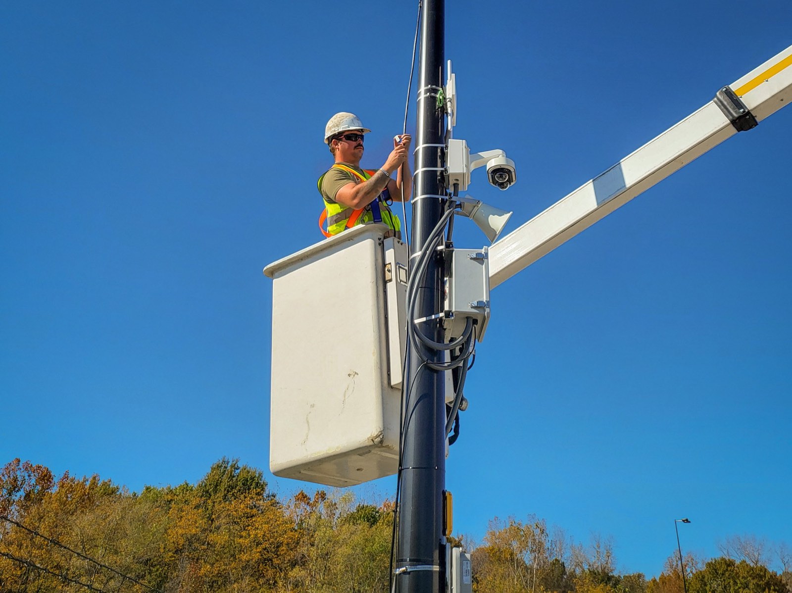 Golden Lab technician in a hard hat and safety vest working from a bucket lift on a pole with a security camera, radio and horn speaker